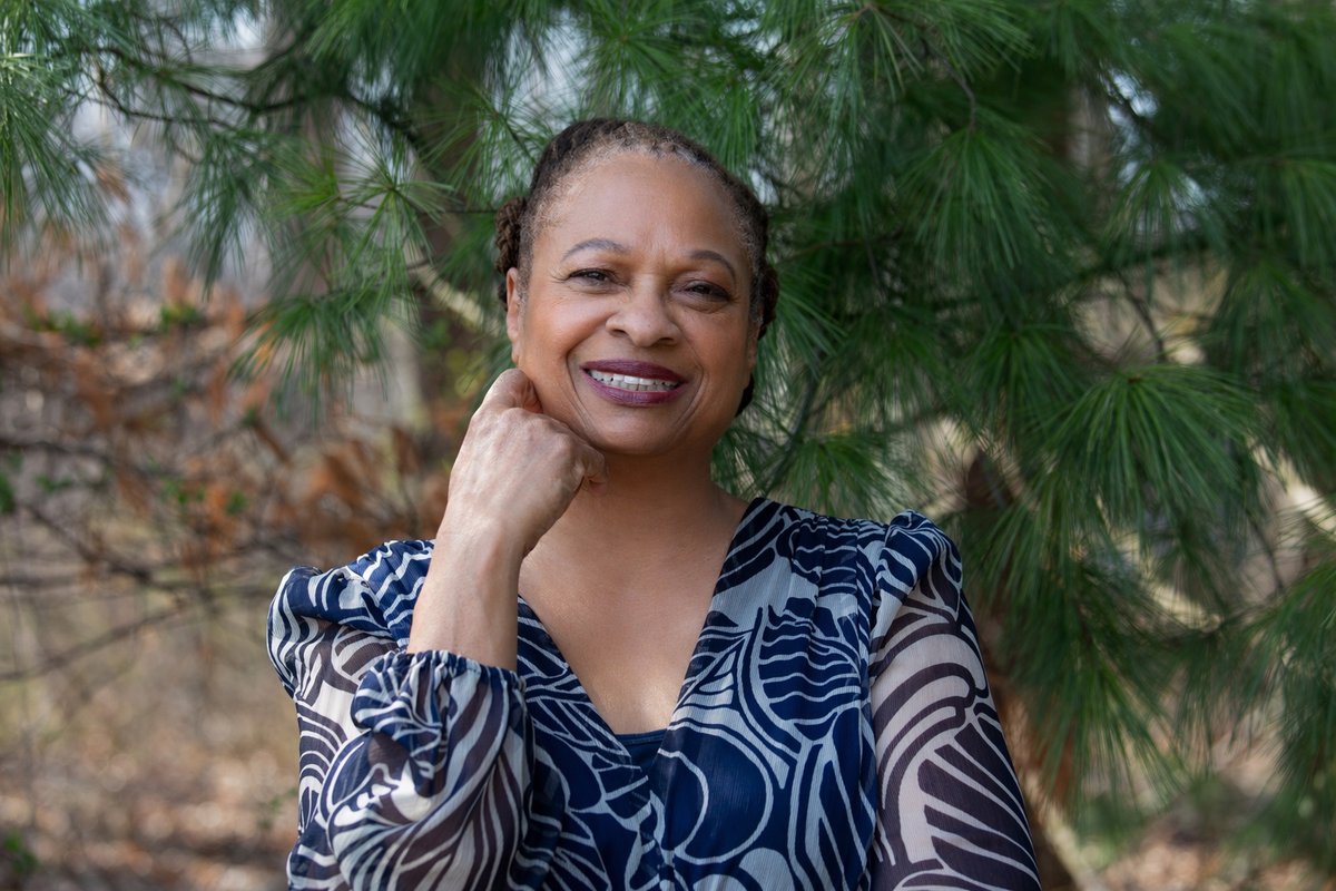 Ruth‑Ann Thompson outdoors among pine branches, smiling toward the camera